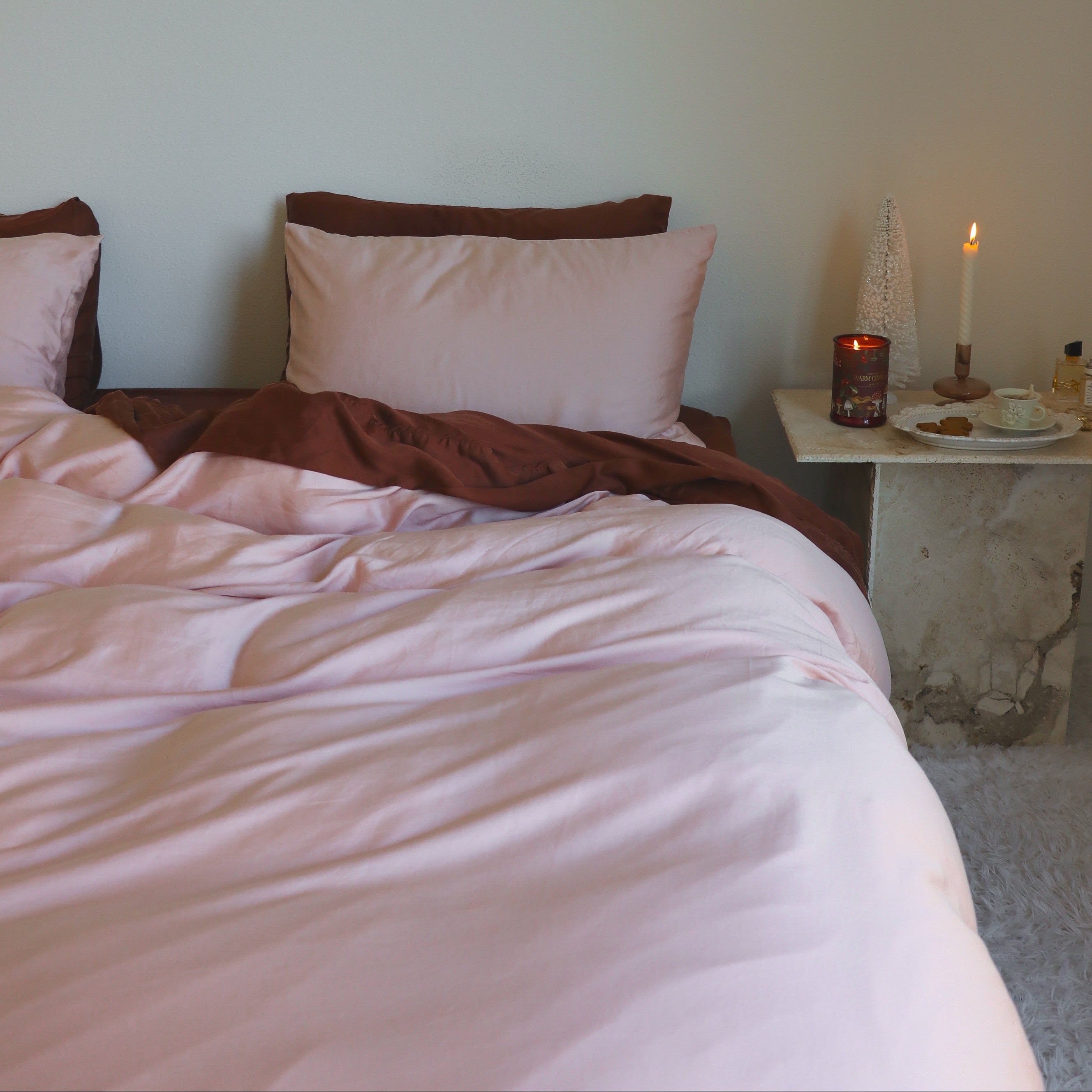Bed with pink bedding and brown blanket, nightstand with candle and small items against a plain wall.